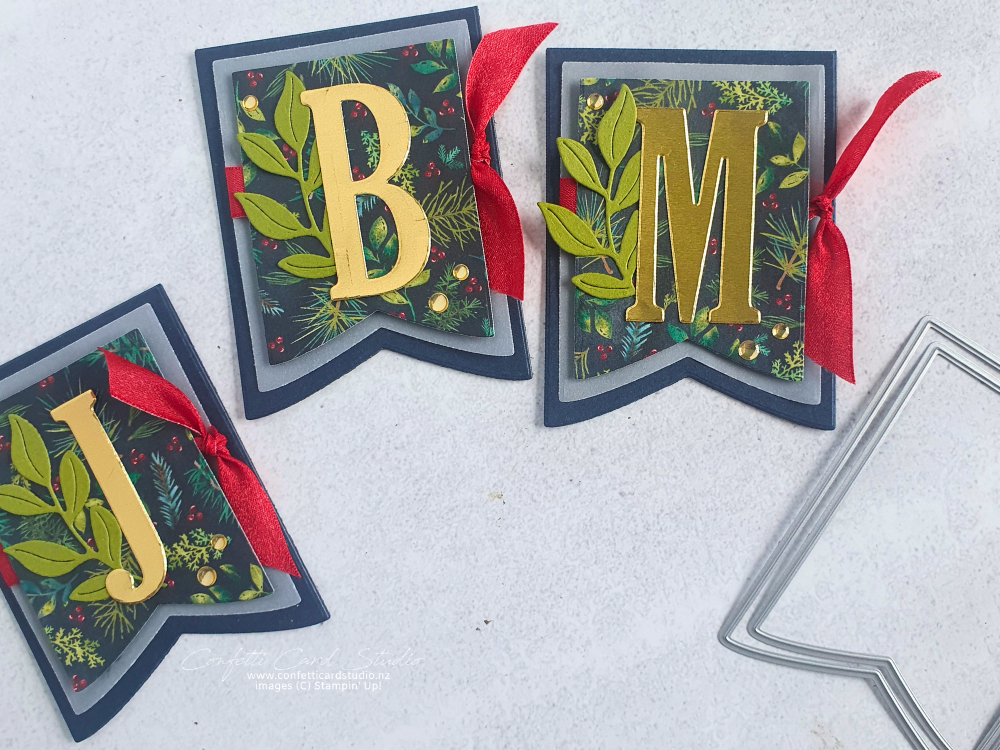 Handmade personalised Christmas table tags. Each dark blue banner shaped tag has an initial in gold. Layers include vellum and pretty green and red festive paper and a red ribbon to the side.