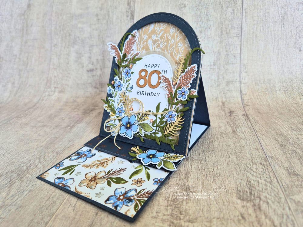 Delicate Whispers Arched Easel Card featuring blue floral layers, vellum arch, bronze accents and an 80th birthday sentiment on a raised easel base.