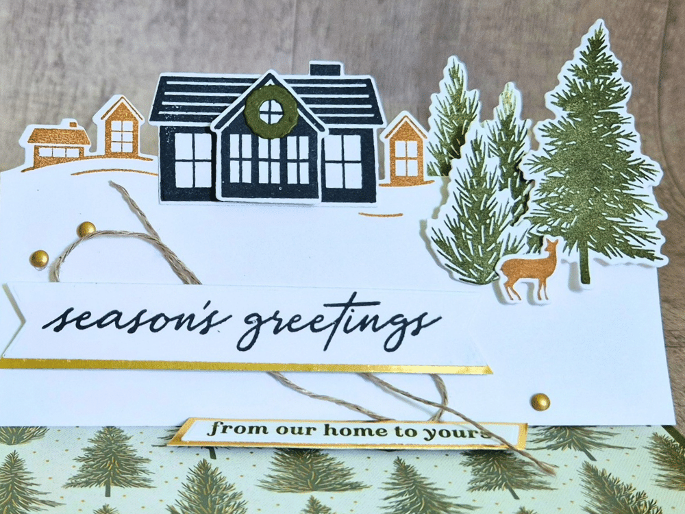 Close up of Stampin’ Up! Welcome Noel easel Christmas card with a village scene, evergreen trees, woodland deer, gold accents and a Season’s Greetings banner.